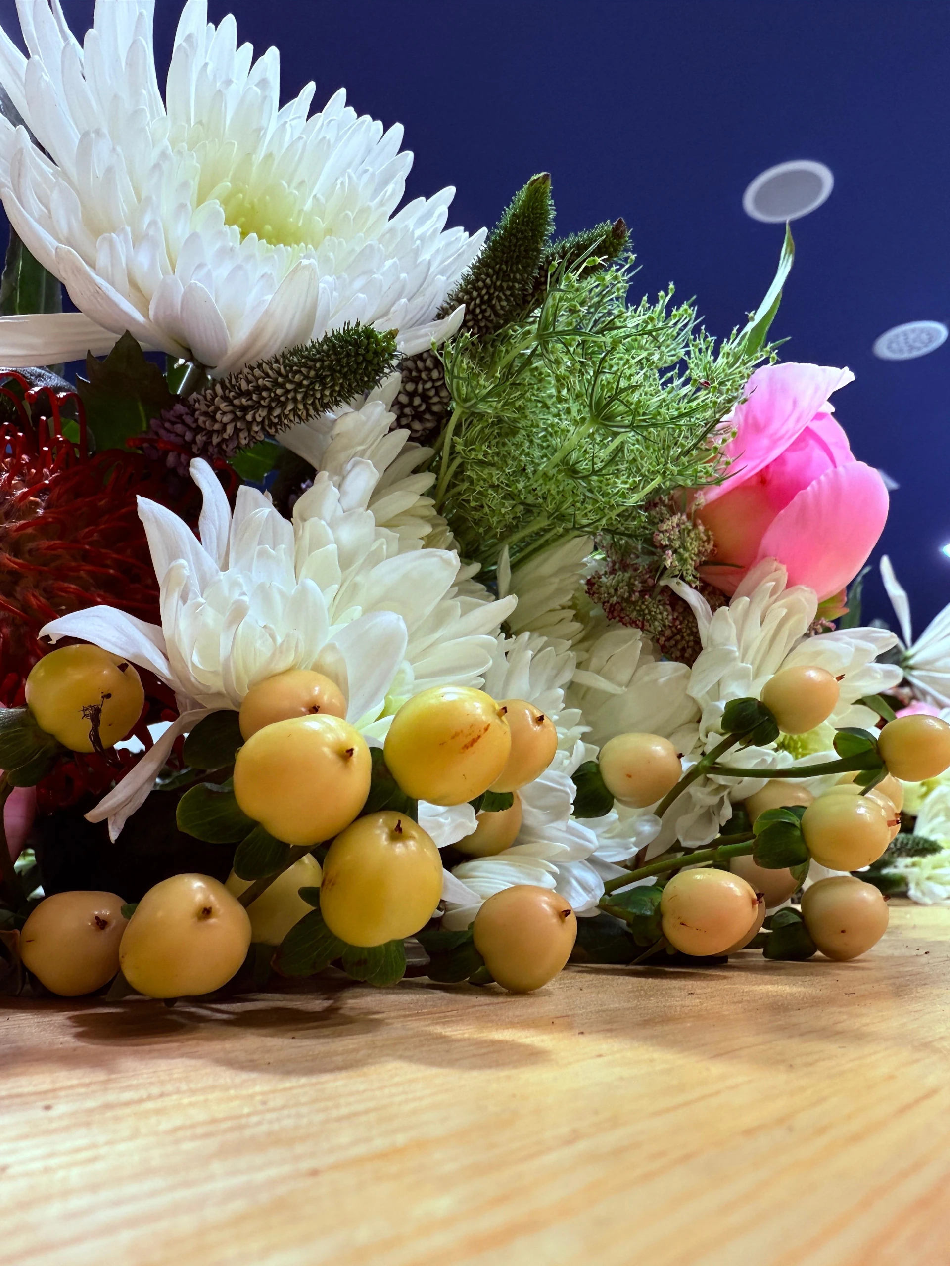 Ram artesanal amb crisantems blancs, roses rosa pàl·lid, fruits grocs i verds frescos sobre fons blau fosc. Una combinació d’elegància natural i contrastos que transmet harmonia i autenticitat.